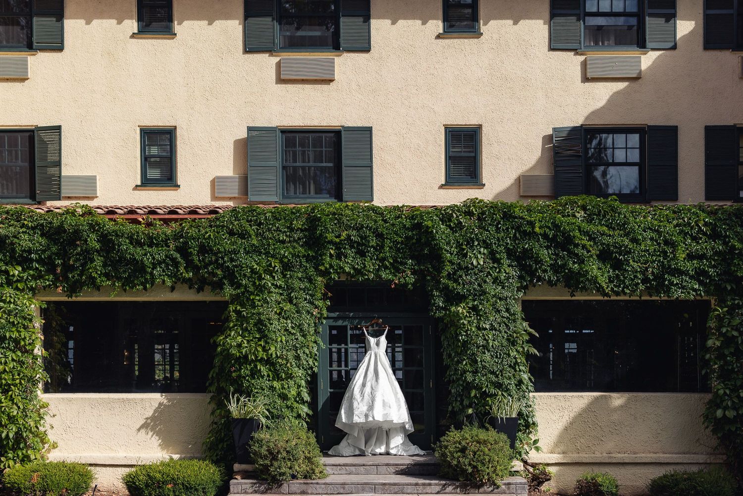 A white wedding dress hangs on display between ivy-covered walls of a cream colored building.