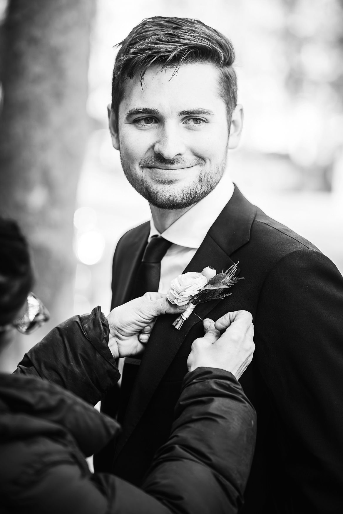 Black and white photo of a groom smiling while someone adjusts his boutonniere before the wedding ceremony.