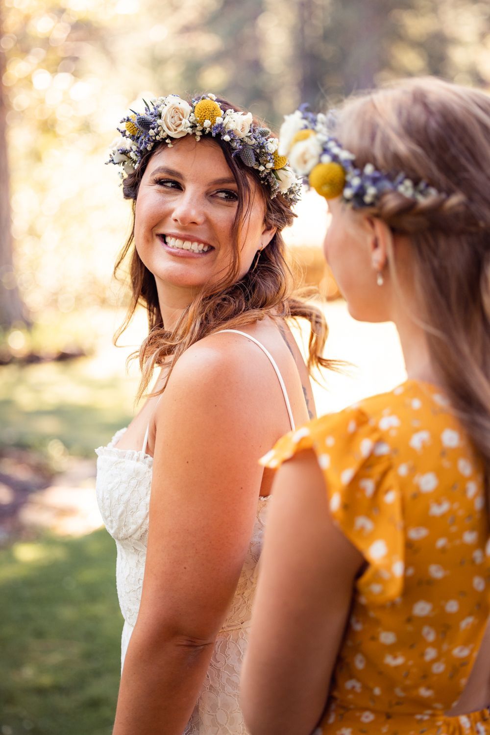 Two women wearing floral crowns and summer attire smile outdoors in natural lighting with a soft bokeh background.