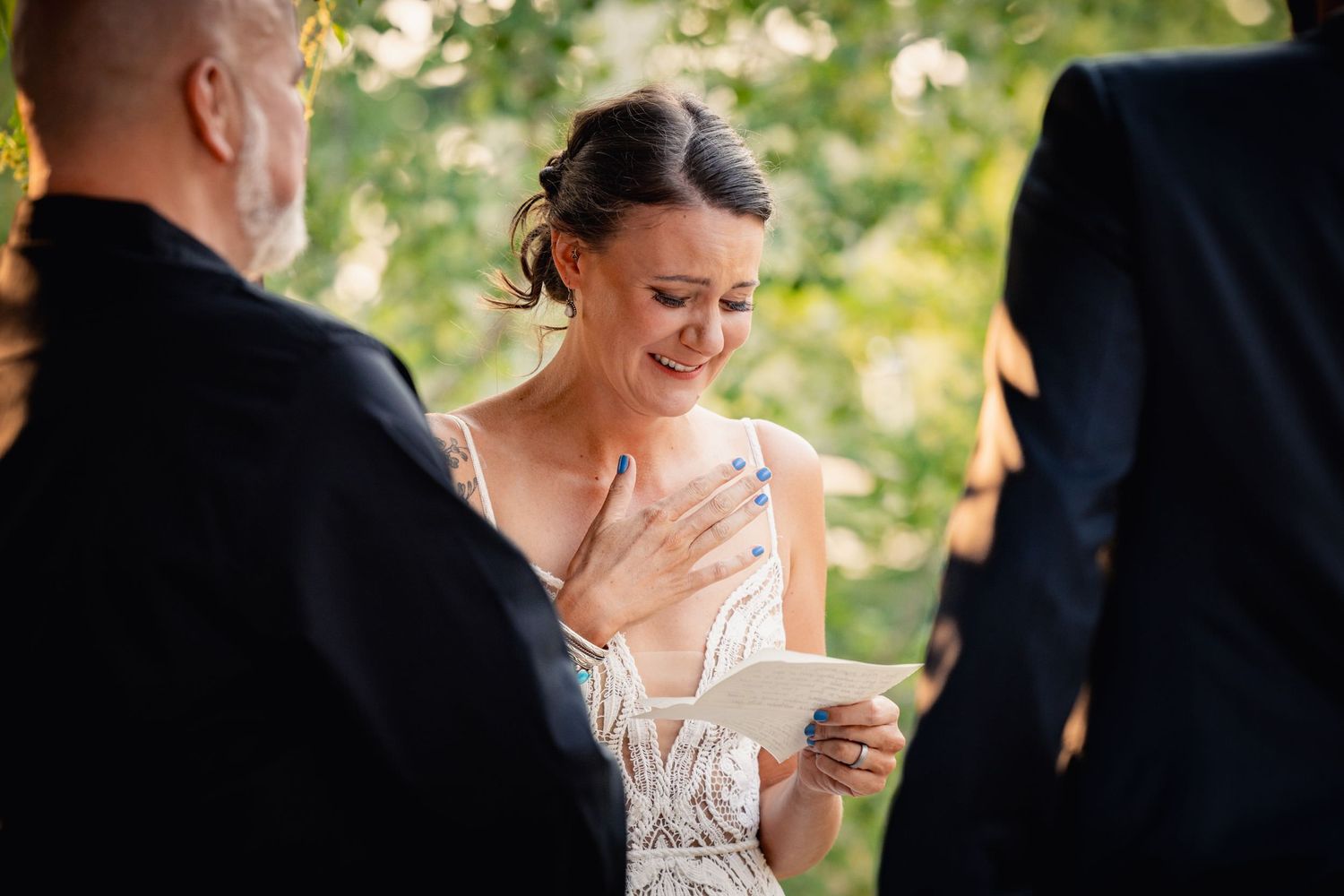 A emotional moment during an outdoor wedding ceremony as the bride reads personal vows among greenery.