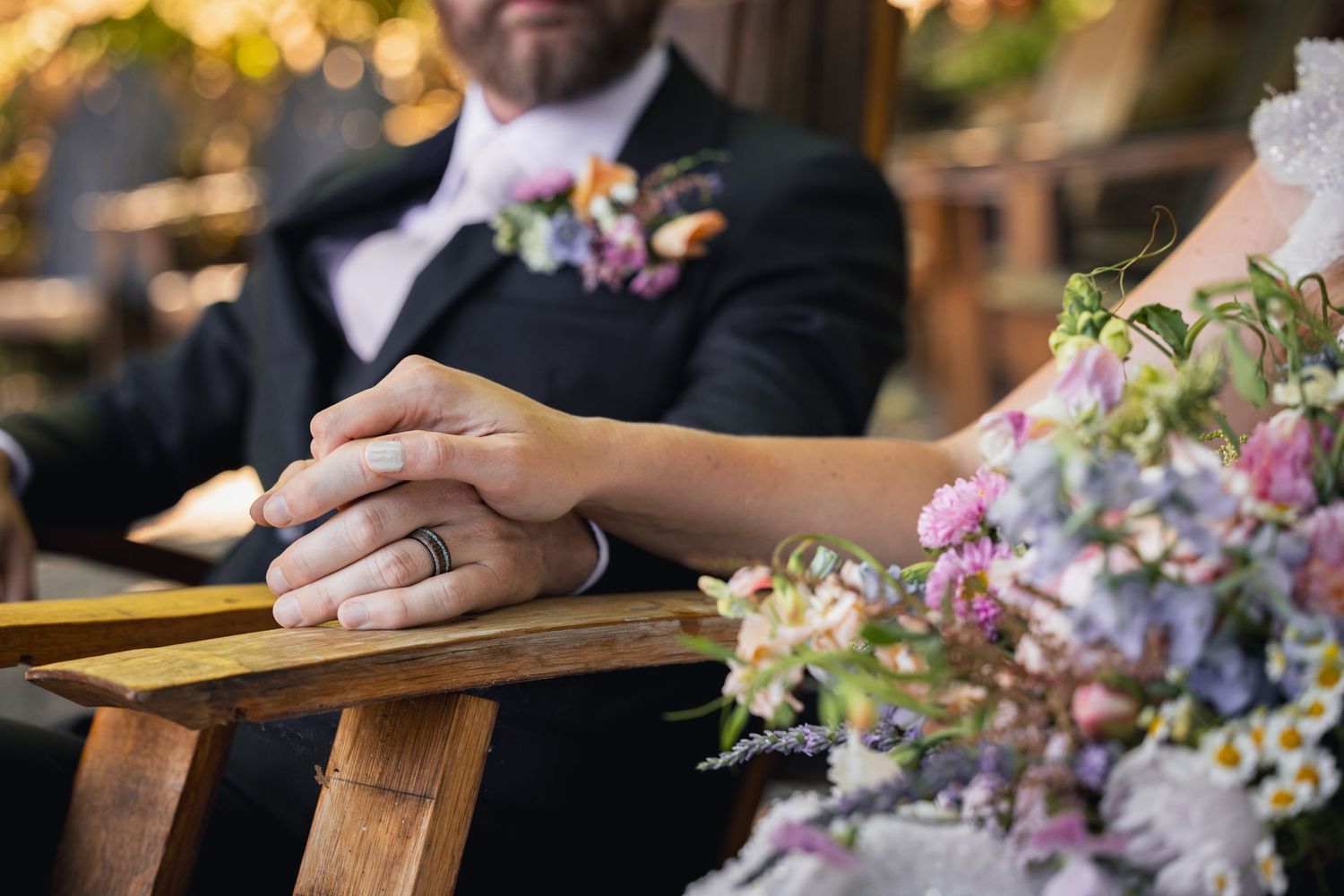 A closeup of clasped hands resting on a wooden surface next to a pastel floral bouquet at a wedding ceremony.