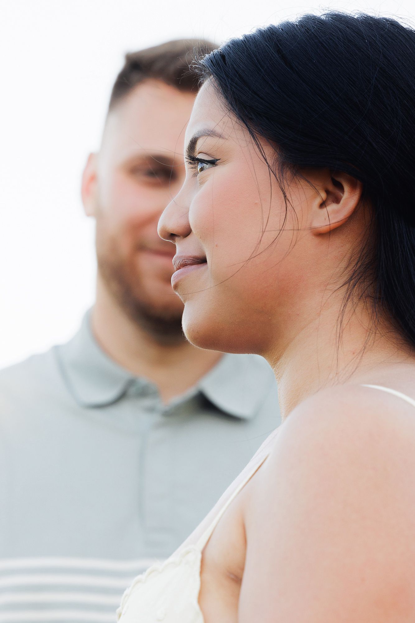 A woman smiles softly in the foreground while a man in a gray polo shirt stands behind her outdoors.