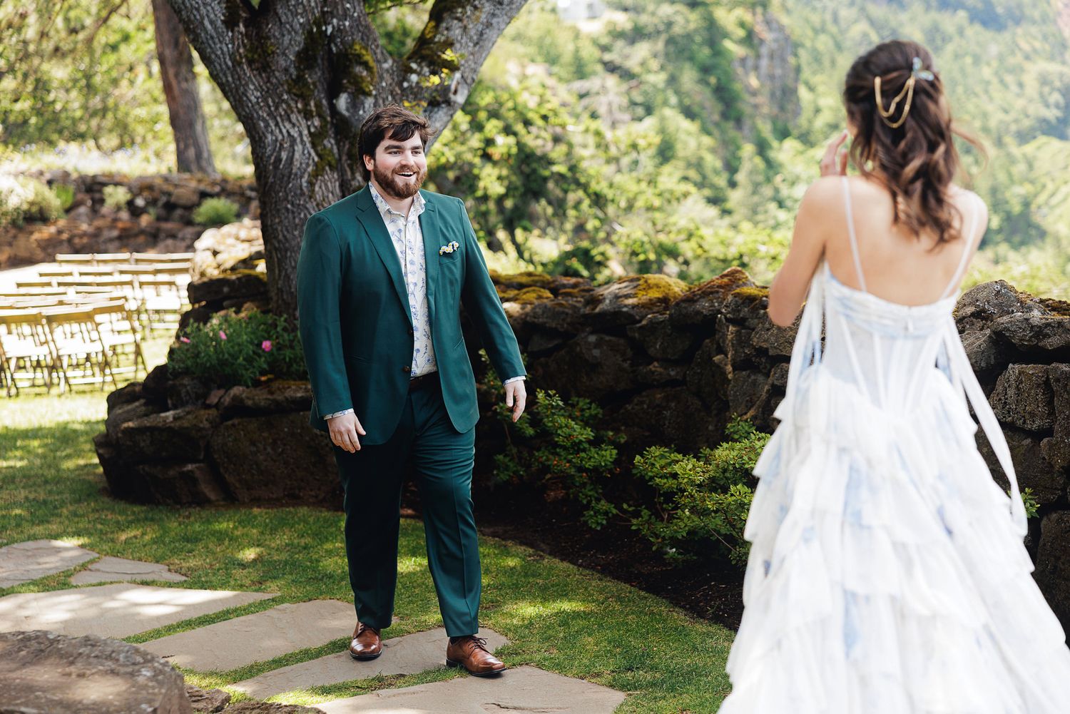 Groom in green suit sees bride in layered white gown for first look at outdoor wedding venue.