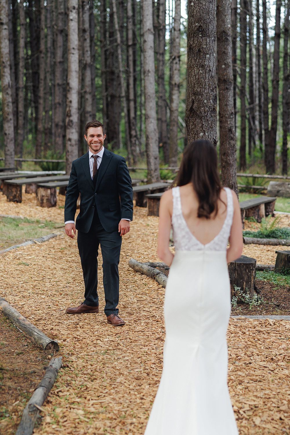 A couple in wedding attire stands apart on a forest path covered in fallen pine needles.
