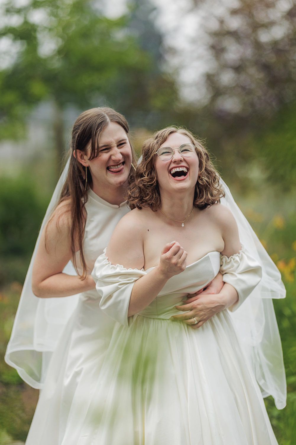 Two people in white dresses share joyful laughter together in an outdoor garden setting.