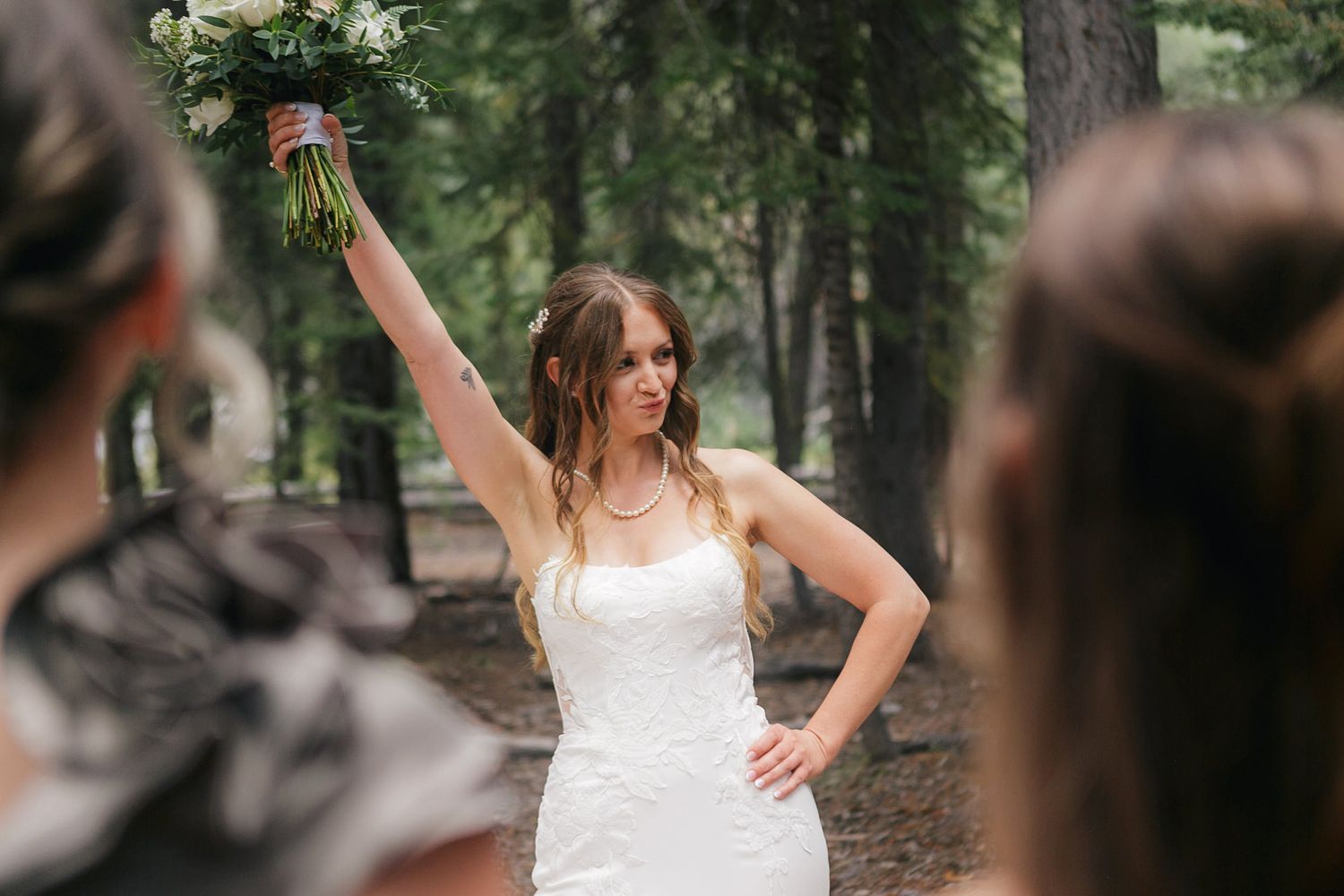 A group of women in white dresses laugh and celebrate together in a wooded outdoor setting.