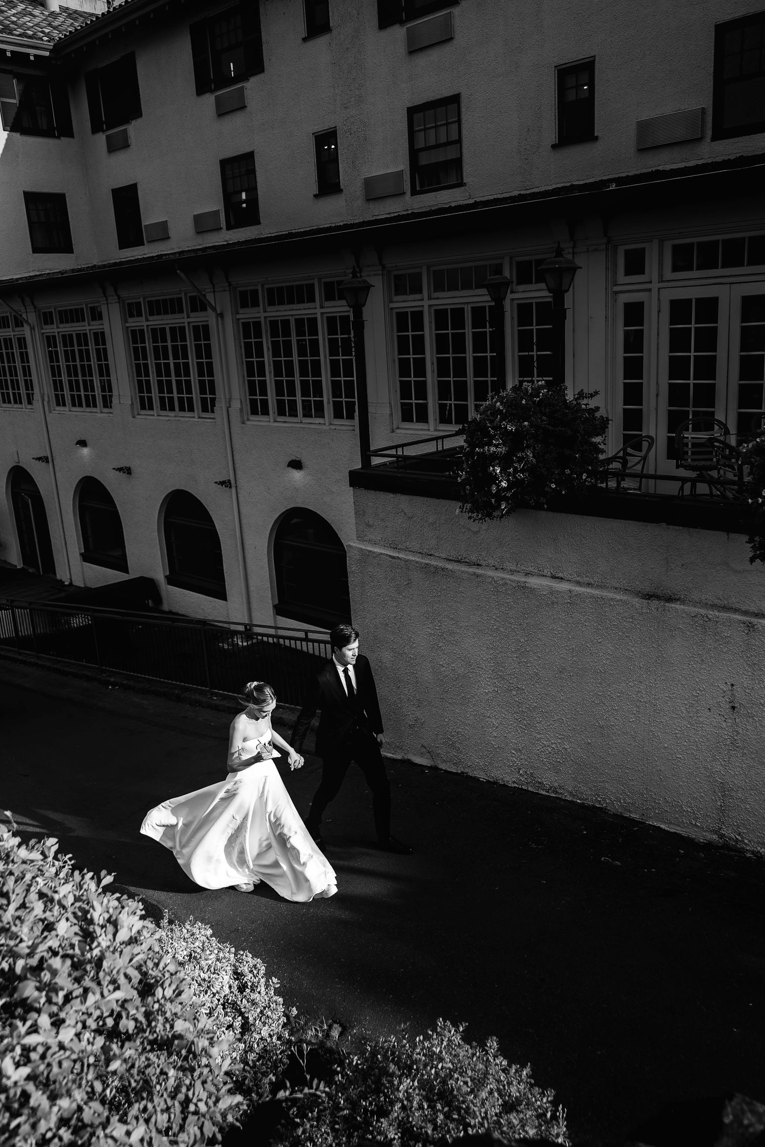 Black and white aerial photo of bride and groom walking outside elegant building, wedding dress flowing dramatically.