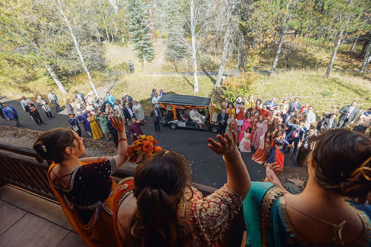 Aerial view of colorful Indian wedding celebration with guests in traditional attire gathered outdoors among autumn trees.