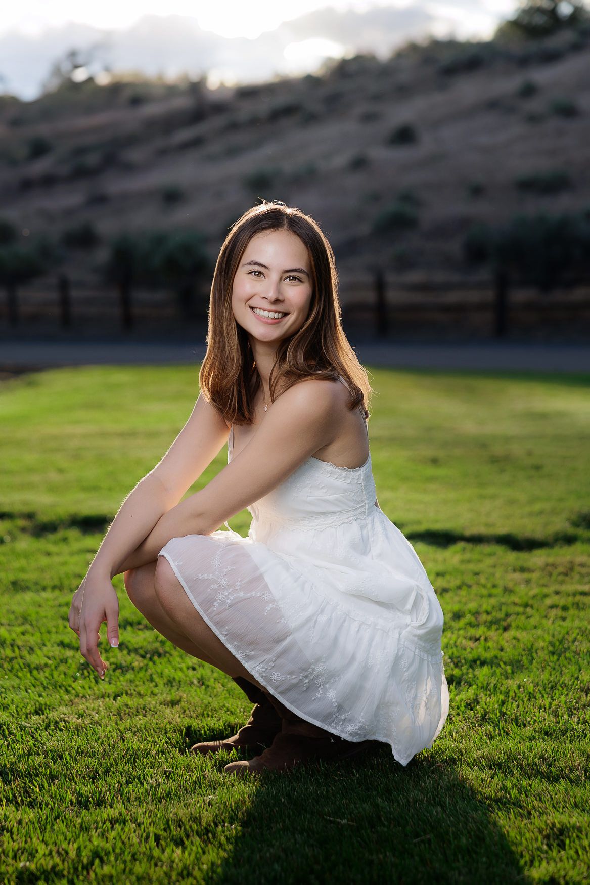 A woman in a flowy white sundress crouches on vibrant green grass with sunlight creating a warm summer glow.
