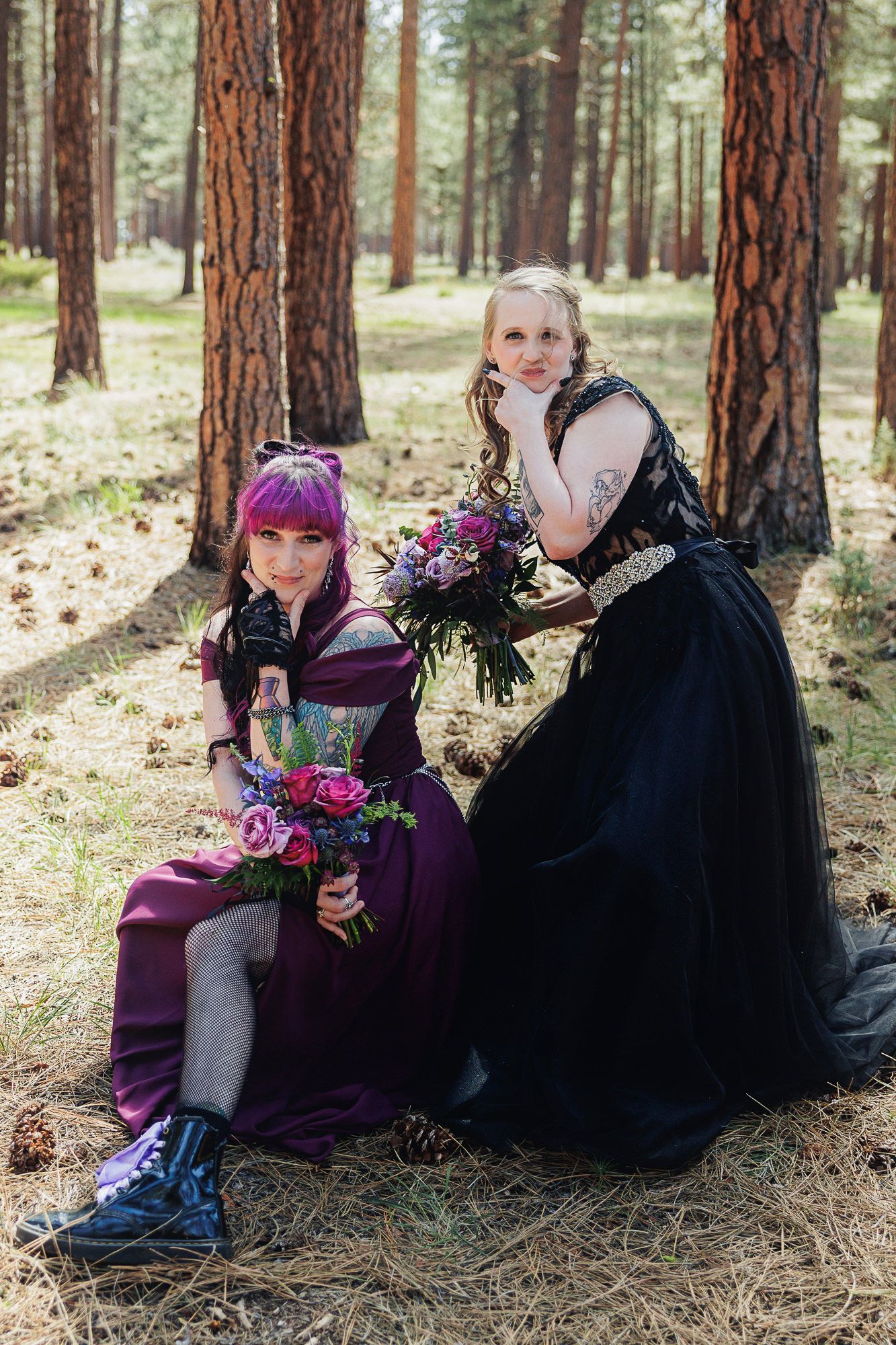 Two brides in purple and black gowns pose in a pine forest, holding colorful bouquets for their gothic wedding.