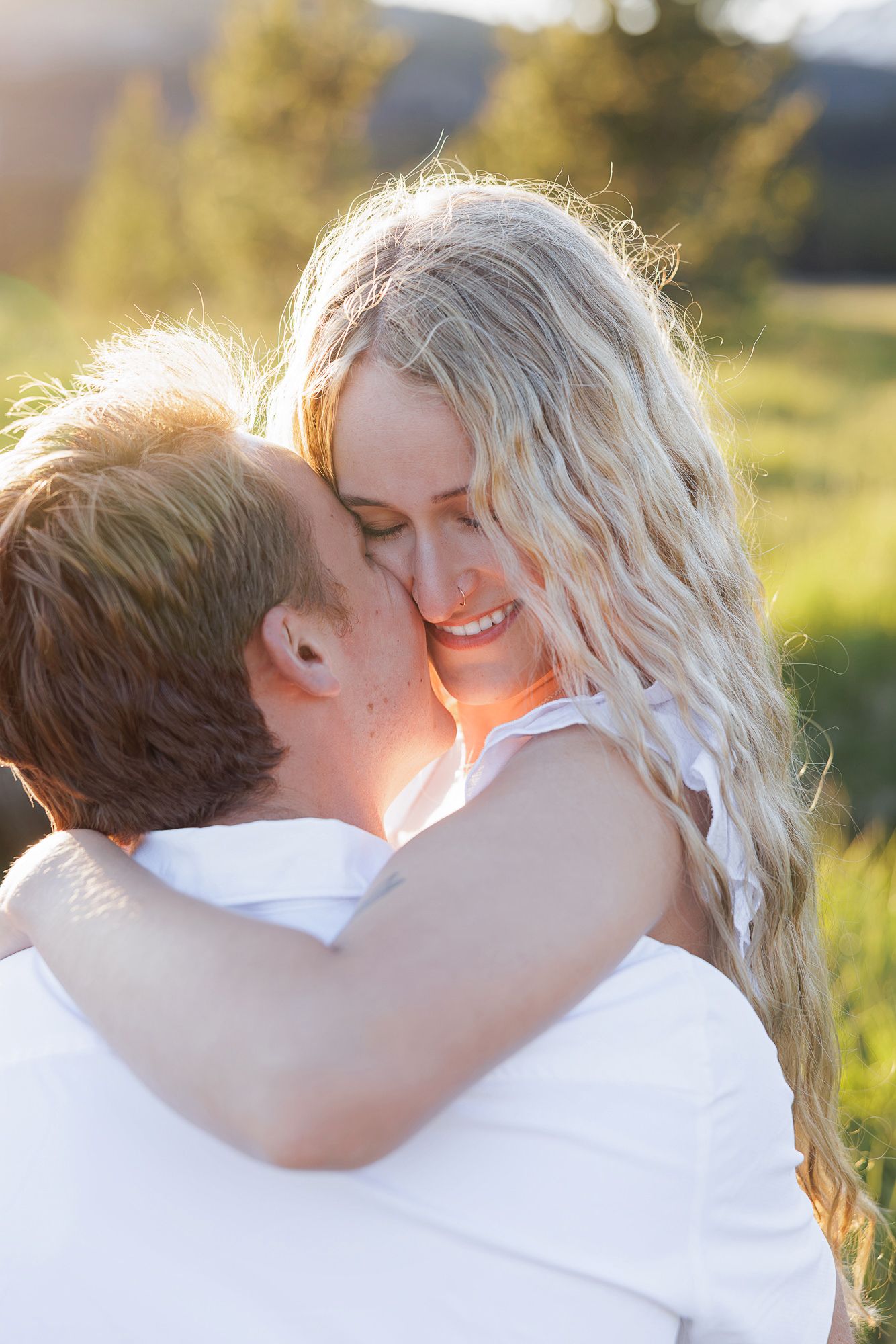 A couple shares a tender moment outdoors, man kissing woman's cheek as she smiles, bathed in golden sunlight.