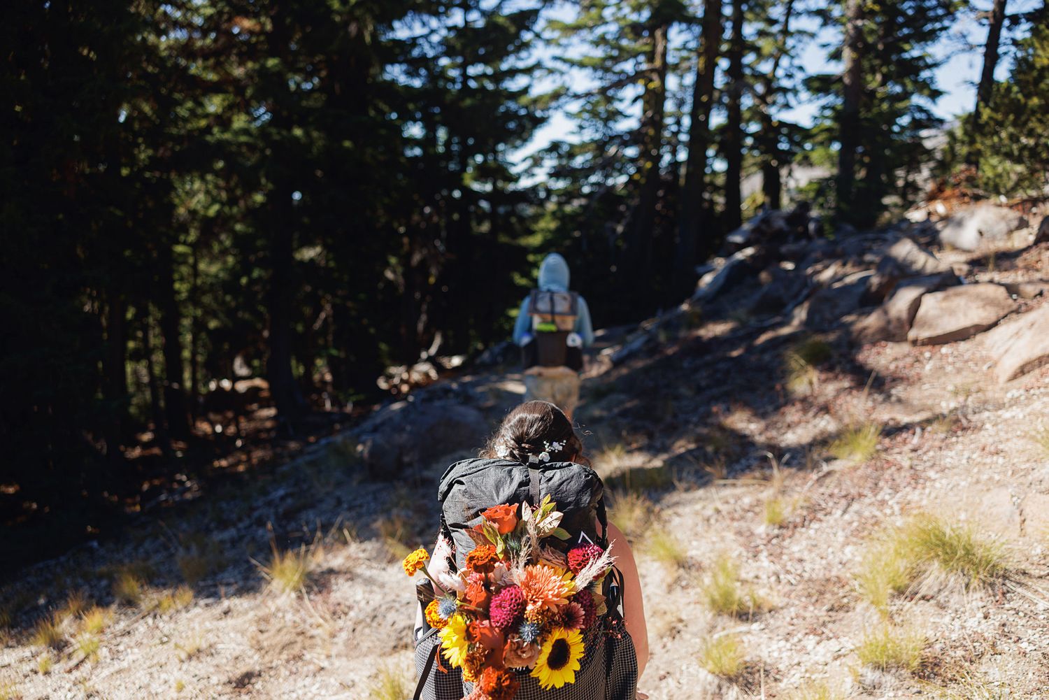 Hiker with a backpack decorated with colorful sunflowers and wildflowers walks a sunlit rocky forest trail while another hiker climbs ahead.