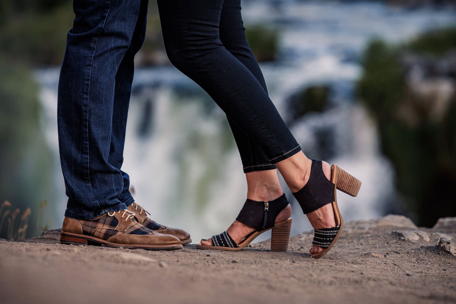 Close-up view of brown leather dress shoes and black heels standing on rocky ground near water.