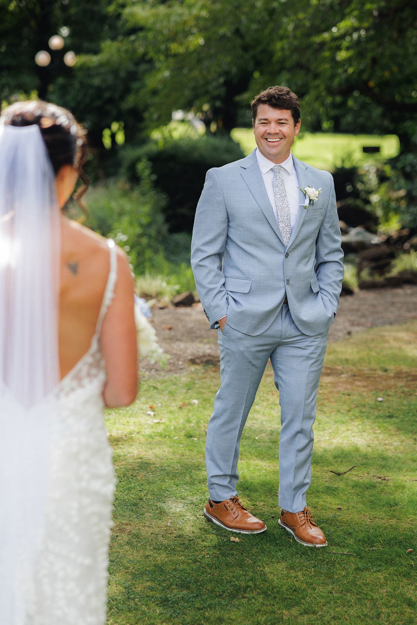 Wedding couple shares playful moments on a grassy lawn in summer with groom wearing light blue suit and brown shoes.