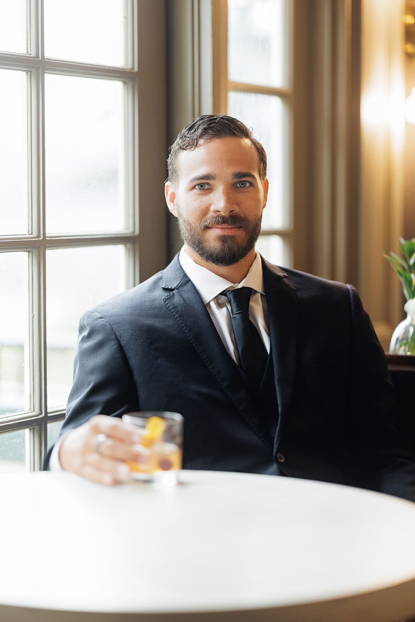 Bearded businessman in dark suit holding whiskey glass at white table near window in elegant interior.