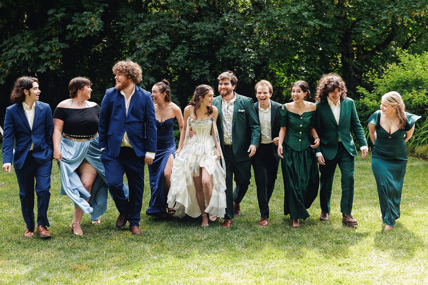 Wedding party walks outdoors in green and blue attire, laughing together on a sunny day.
