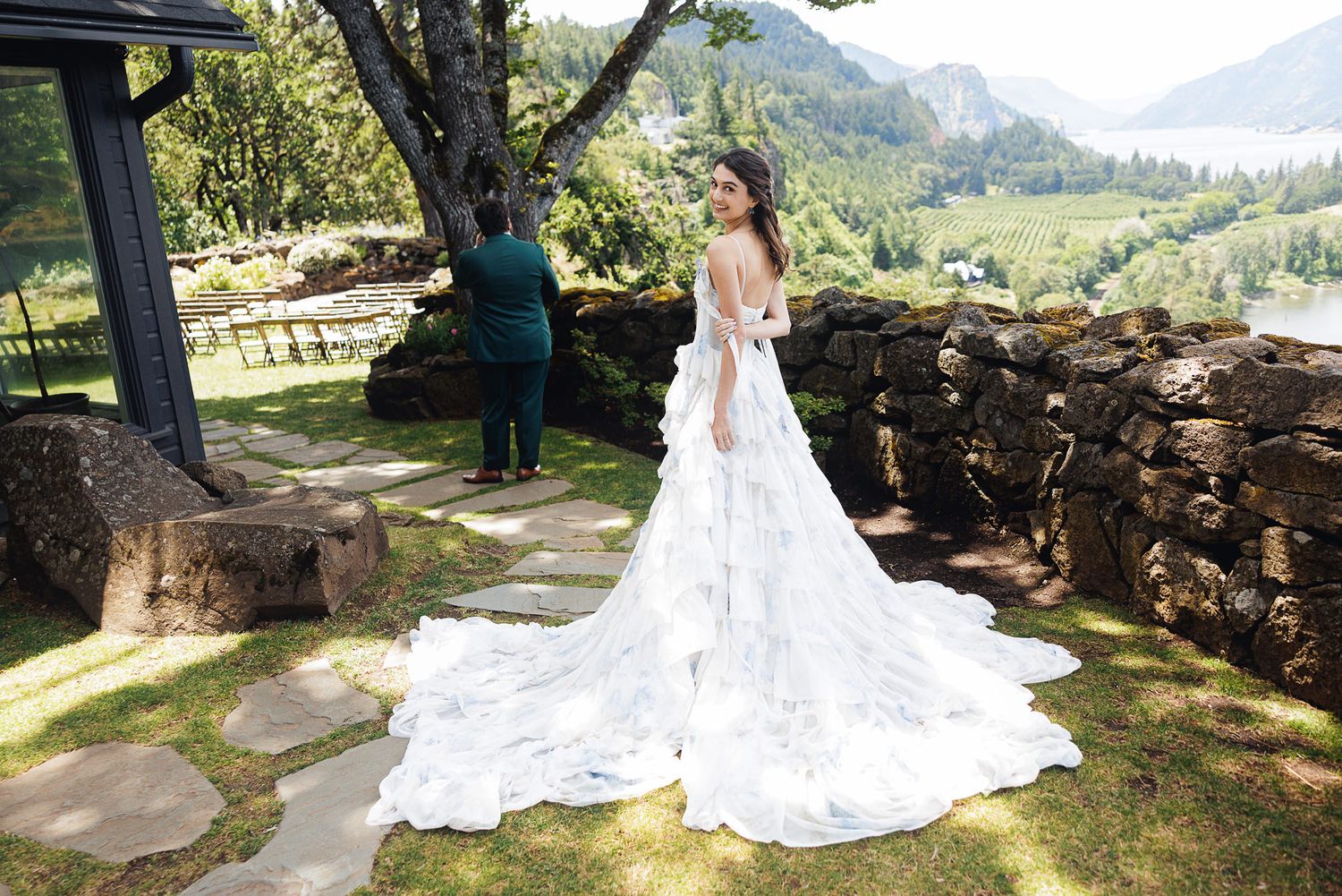 Bride in elegant white ball gown with floral train poses outdoors at scenic mountain venue with stone wall.