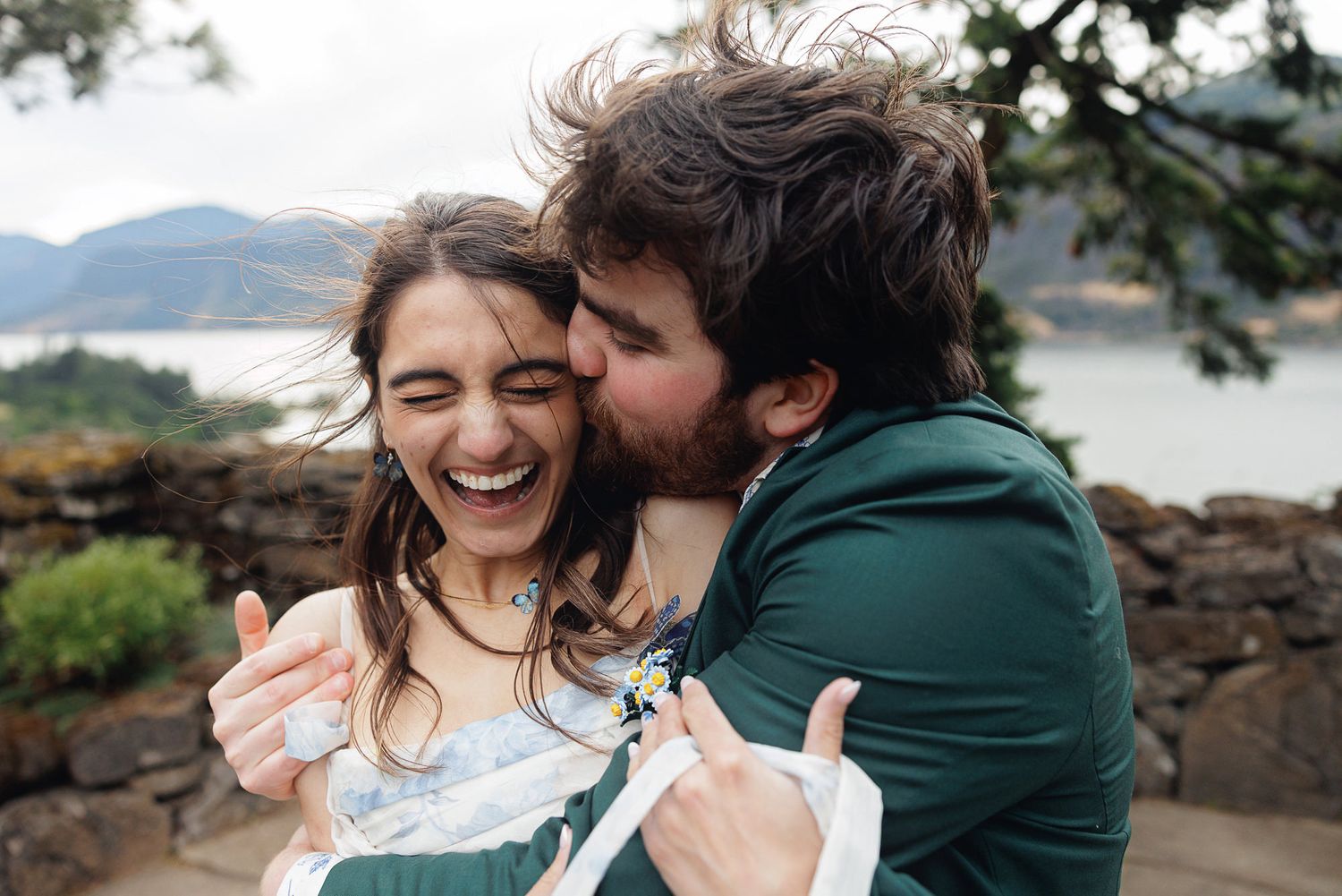Happy couple embracing outdoors, woman laughing joyfully as man kisses her cheek with scenic lake view behind them.