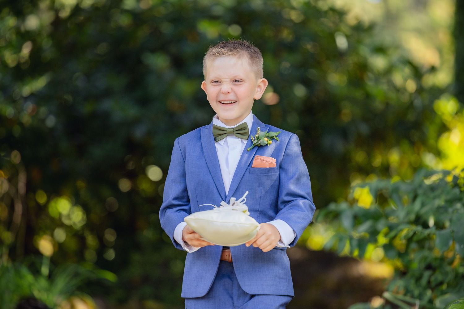 Ring bearer in a blue suit and bowtie smiling while holding a white pillow at an outdoor wedding ceremony.