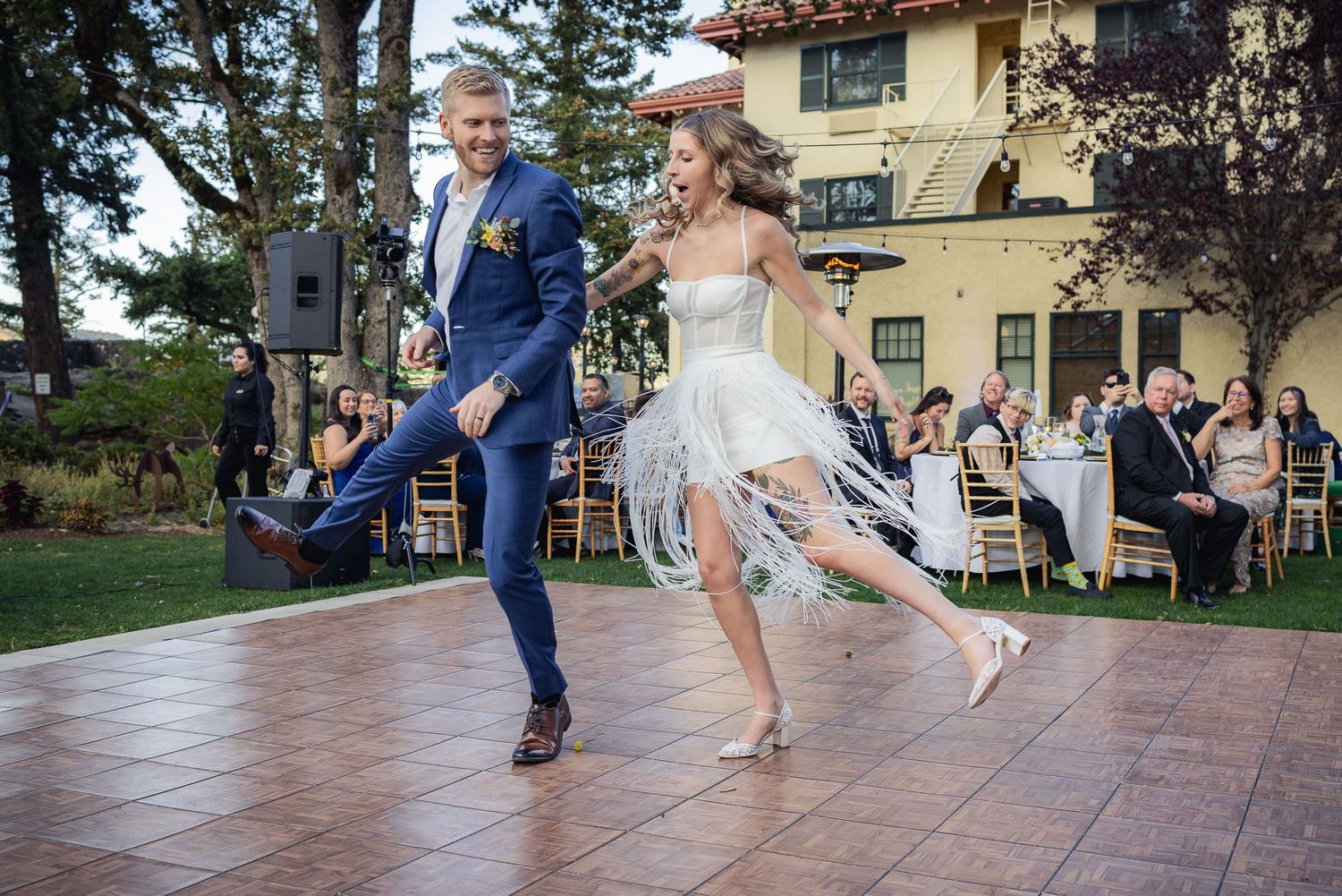 A joyful couple leaps across an outdoor dance floor at their wedding reception with guests seated in the background.