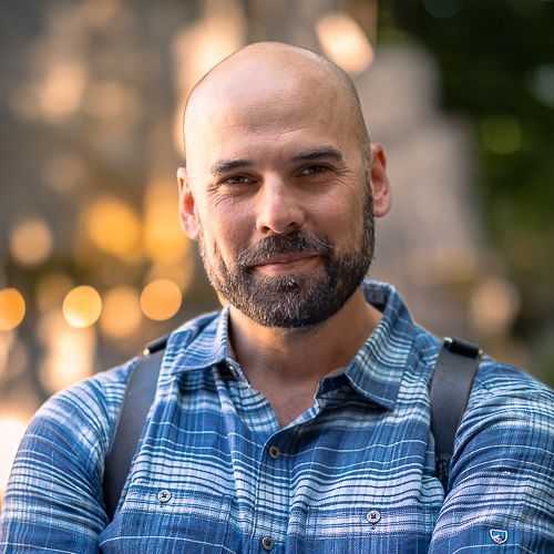 A portrait of a person in a blue plaid shirt smiling at the camera with a blurred bokeh background.
