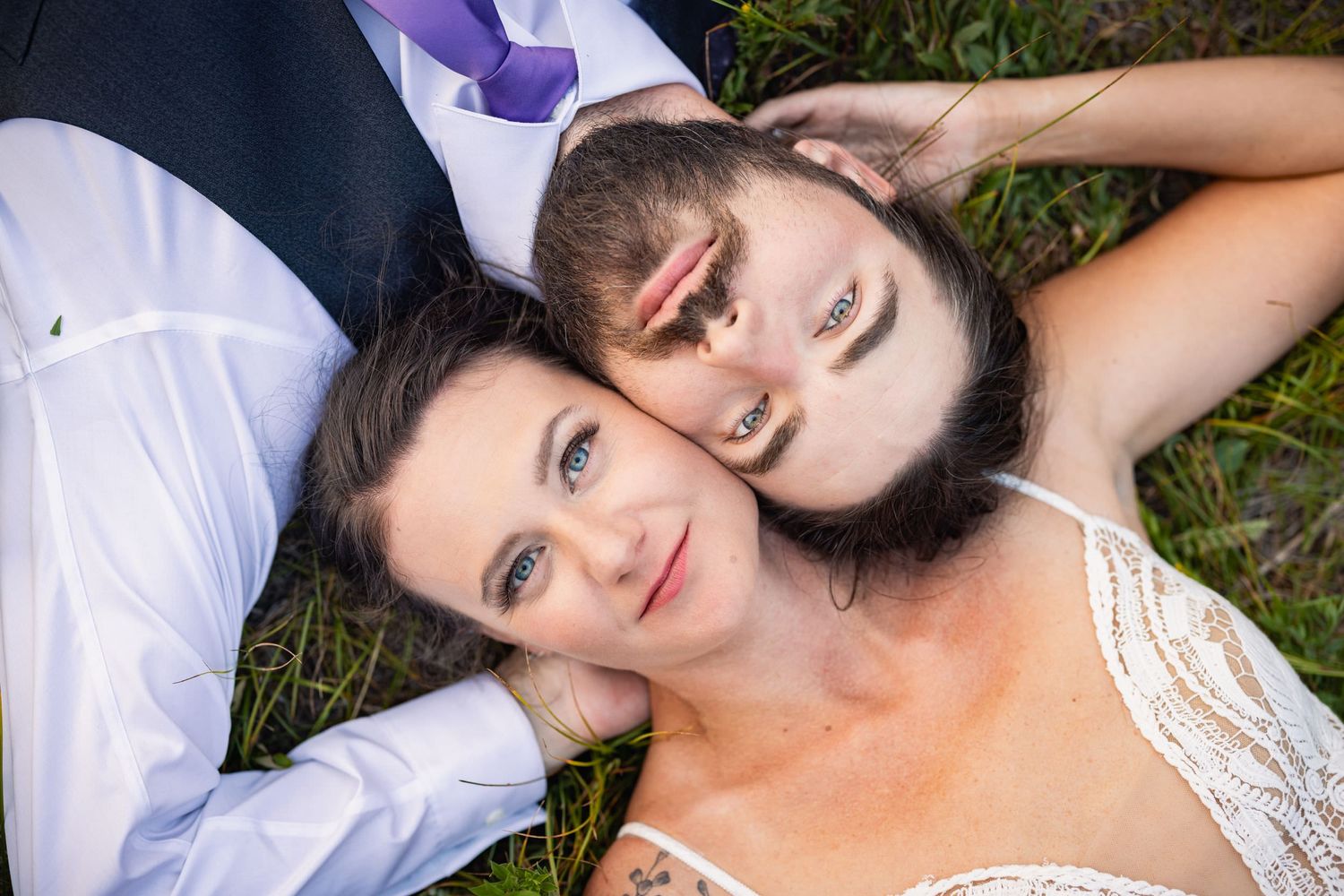 Two people lying on grass wearing formal white attire looking up at camera from above.
