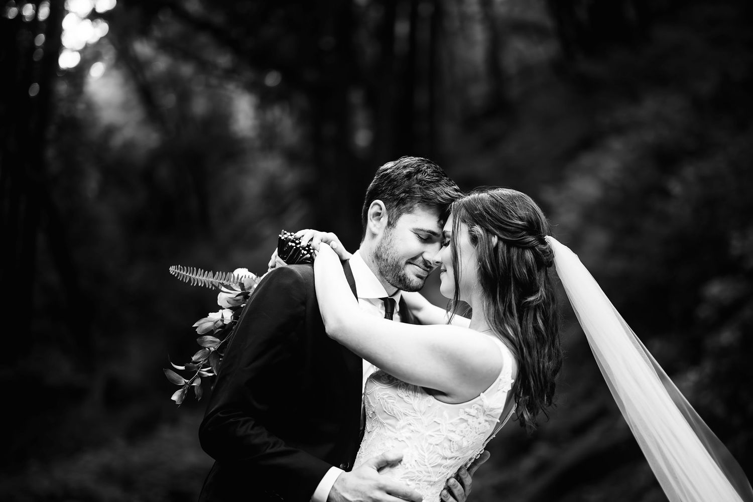 A romantic black and white wedding photo captures an intimate moment between newlyweds in a forest setting.