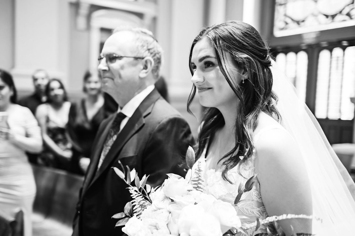 Black and white photo of a bride in a wedding dress holding a bouquet walking down the aisle in a church.