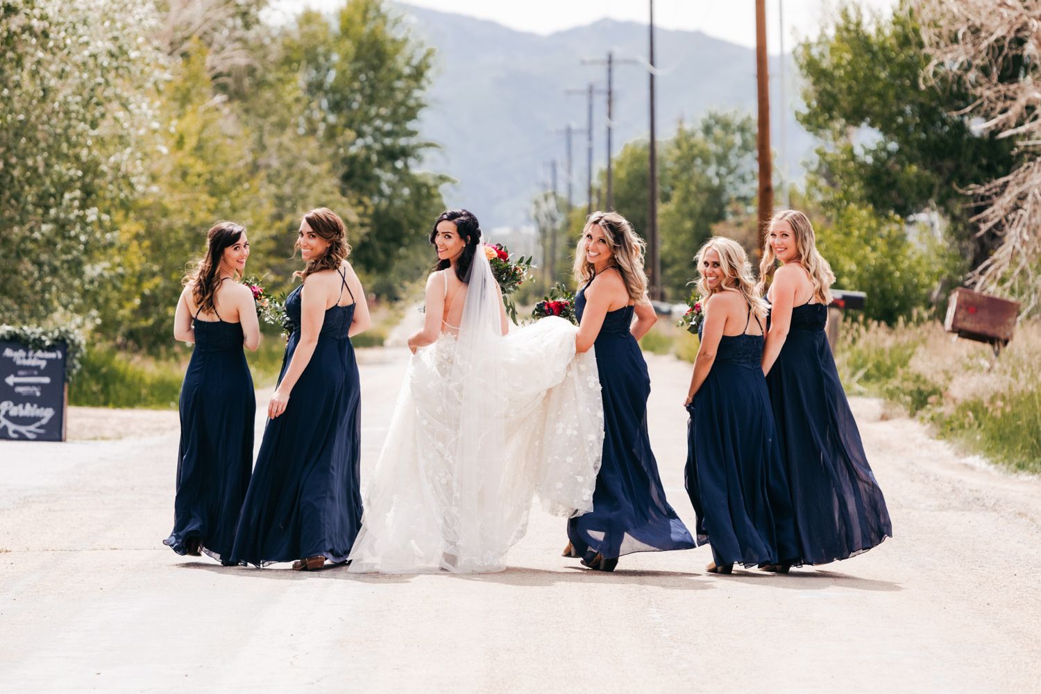 A bridal party in navy blue gowns and white wedding dress walk down a mountain road on a sunny autumn day.