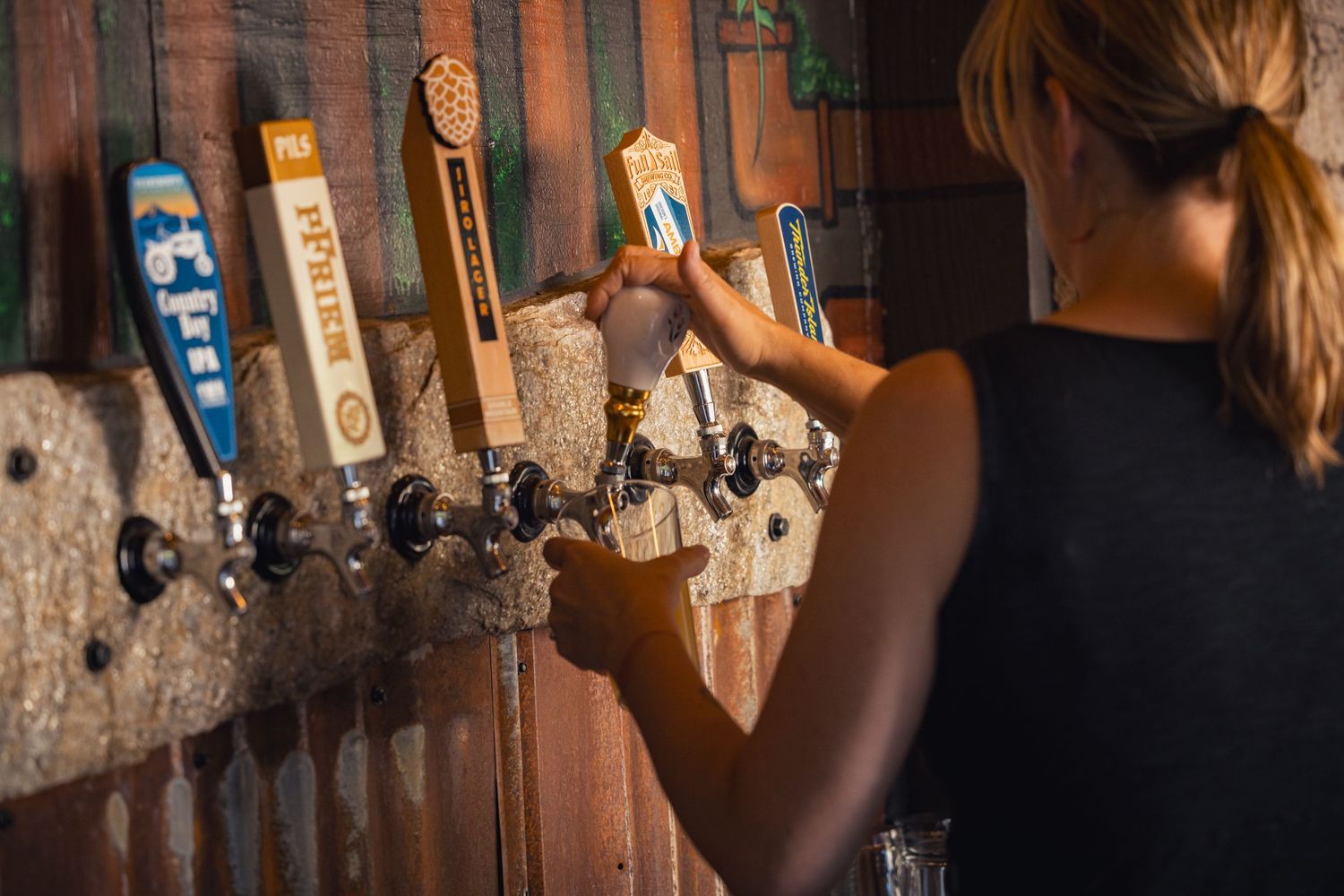 A bartender pours a draft beer from one of several taps mounted on a rustic wooden wall in warm lighting.