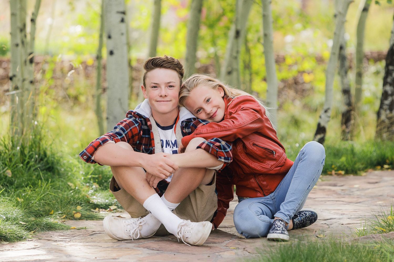 Two young friends sitting casually together on a wooden path through aspen trees.