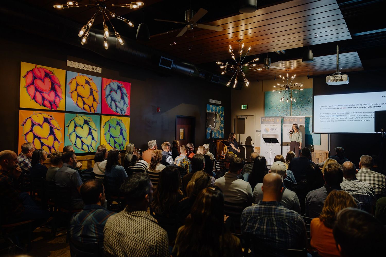 Packed audience watching presentation in dark venue with colorful pop art style wall decor.