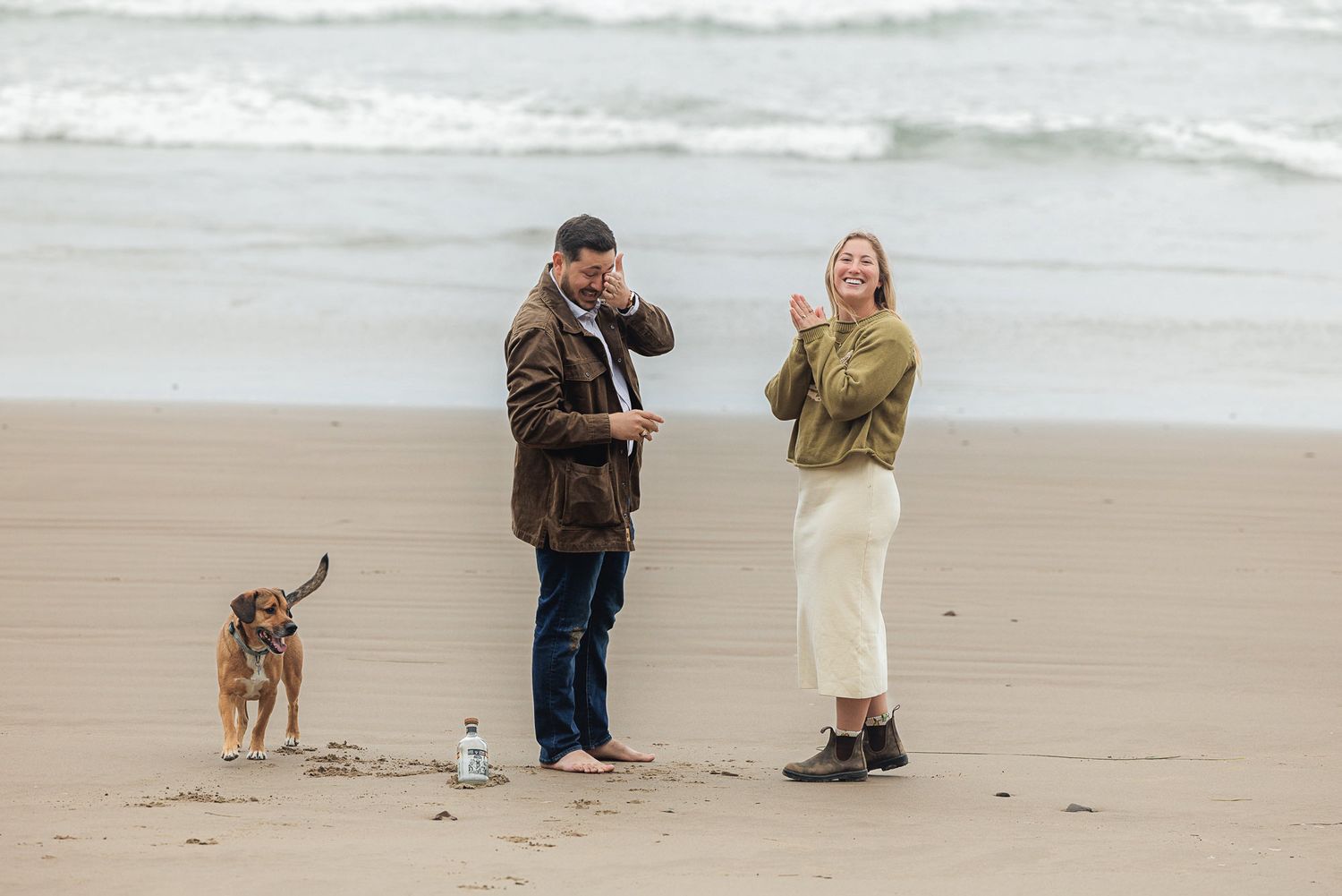 A couple laughs and plays with their dog on a sandy beach, dressed in casual autumn outfits near the ocean waves.