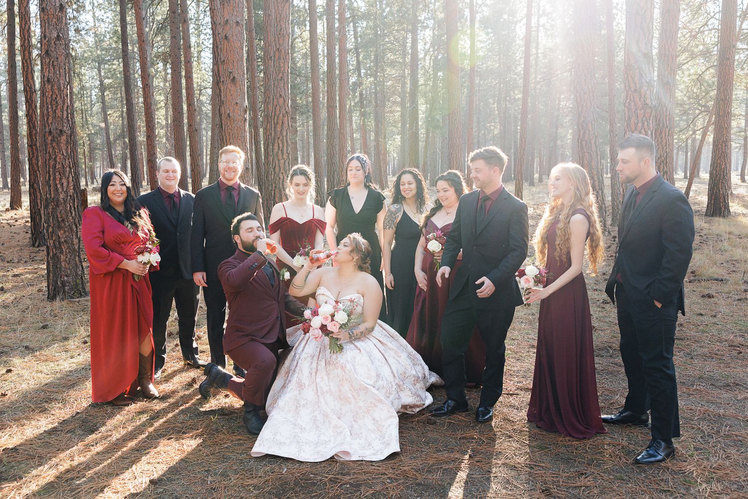 Wedding party in burgundy and black attire poses together in a sunlit forest setting.