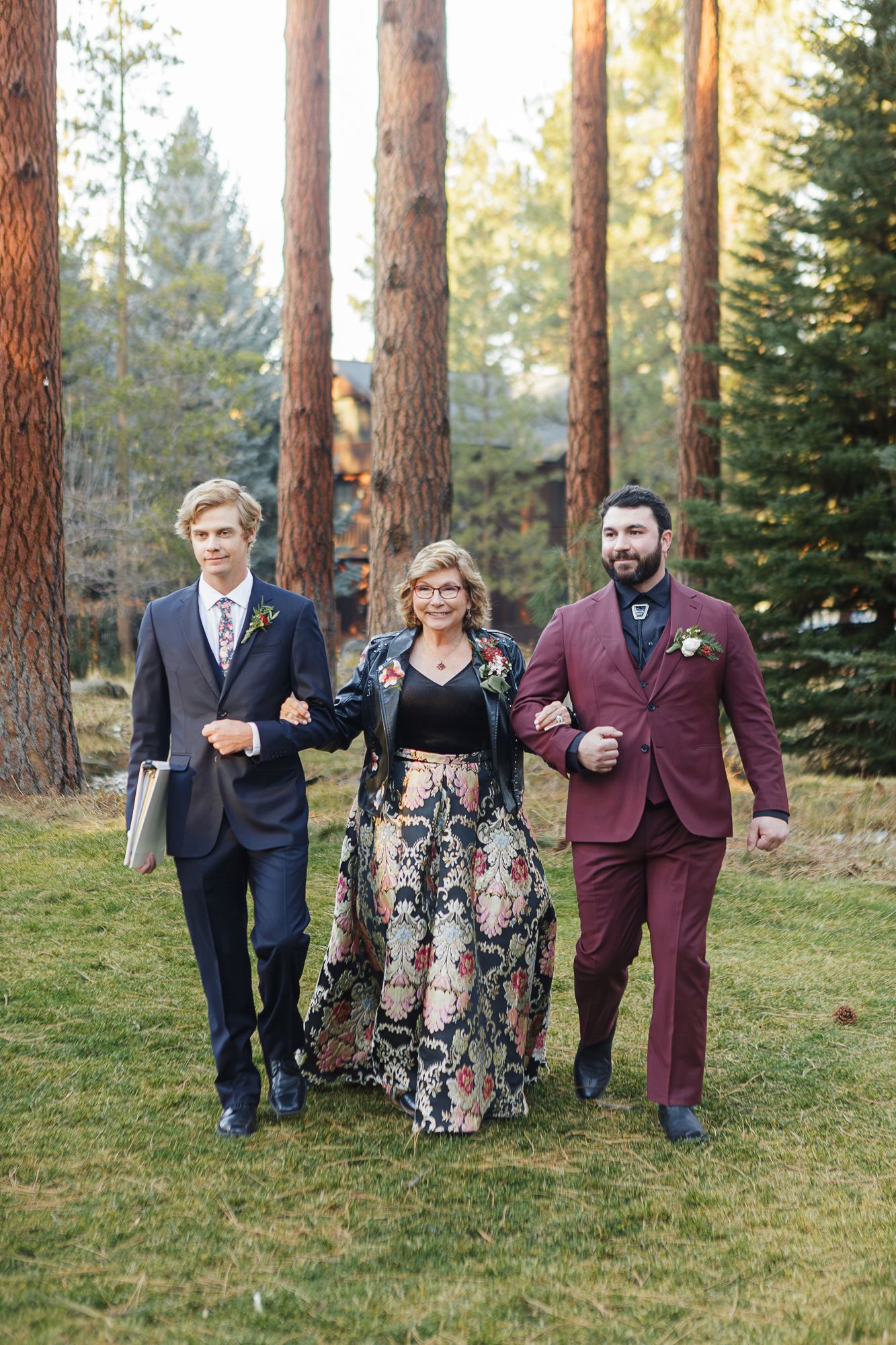 Wedding party in formal attire walks through a forest of tall pine trees on a sunny afternoon.