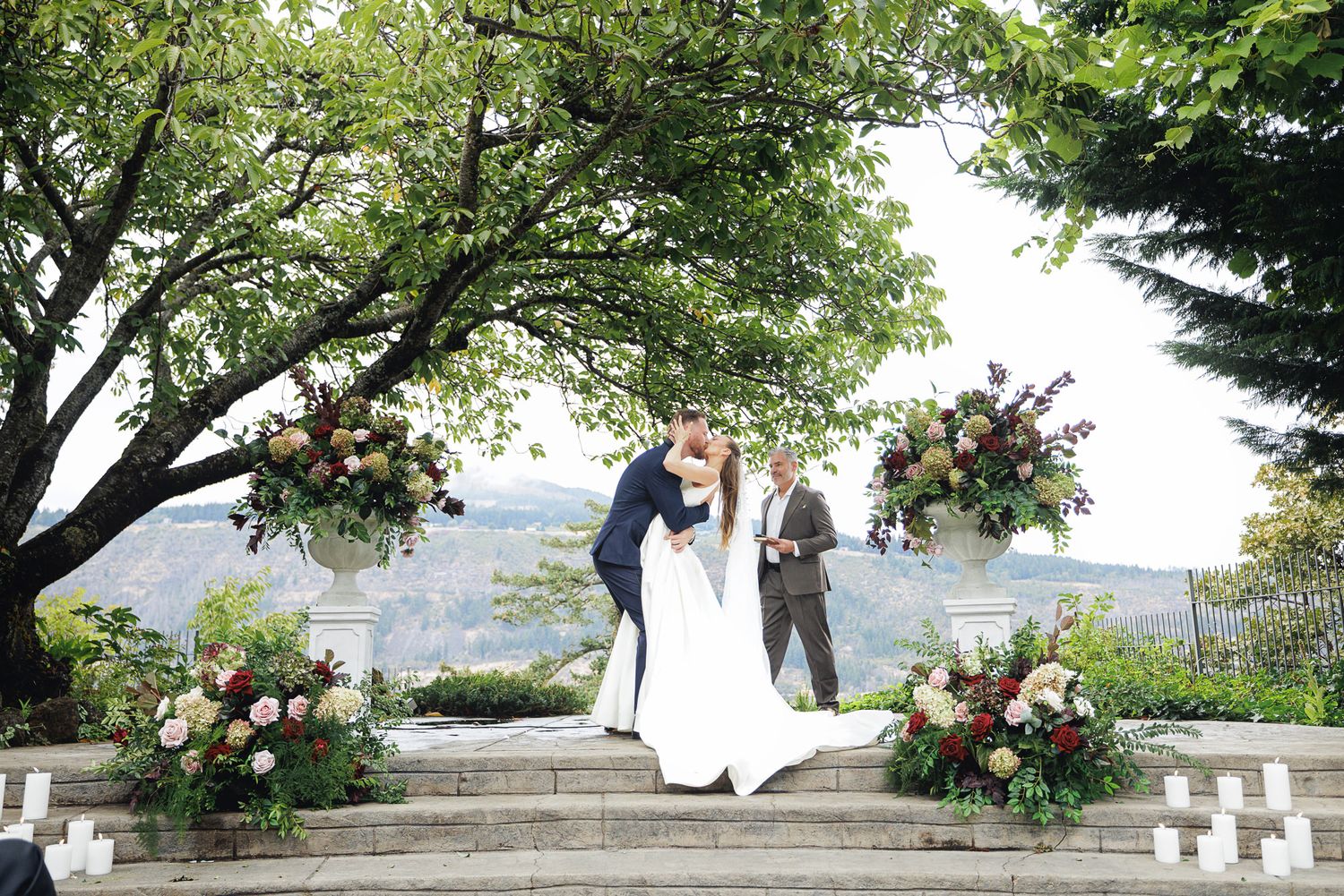 Couple shares first kiss at outdoor wedding ceremony with floral arrangements and scenic mountain backdrop.