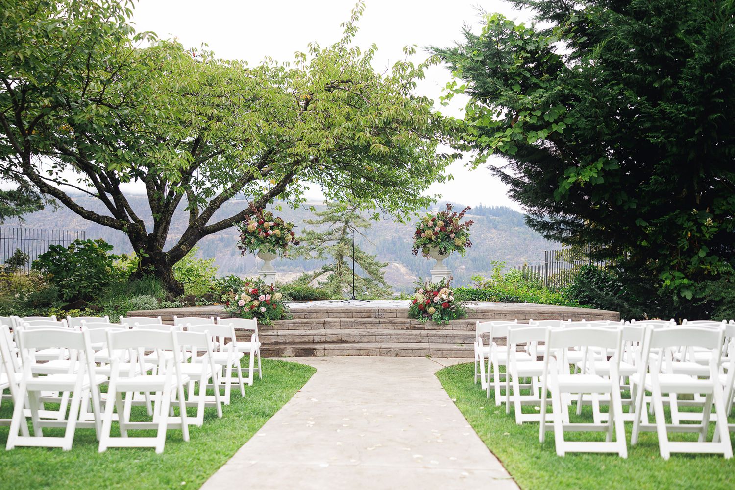Outdoor wedding ceremony setup with white chairs, floral arch, lush greenery, and mountain views in background.