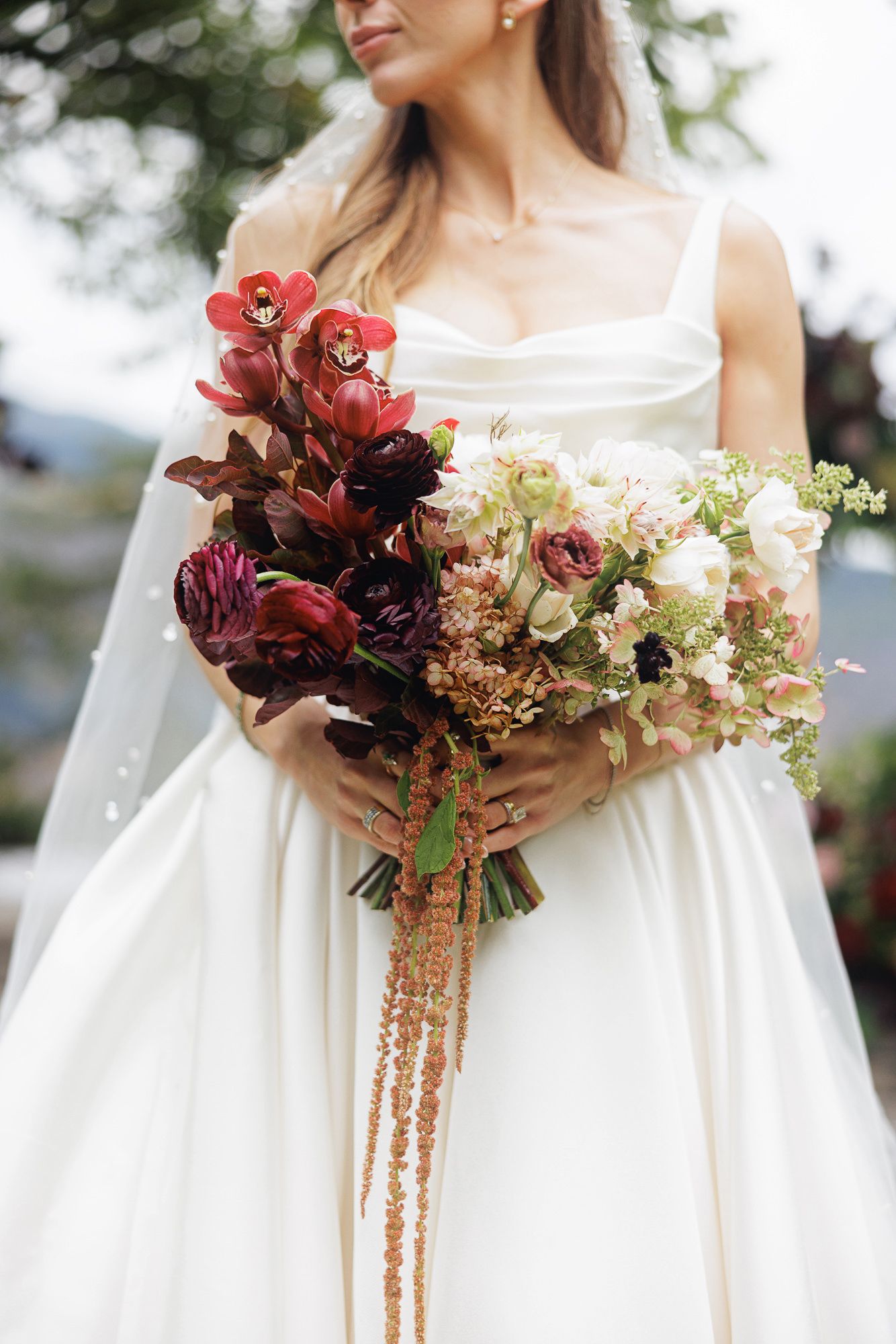 Bride in white gown holding lush bouquet with deep red, burgundy, and blush flowers with trailing accents.