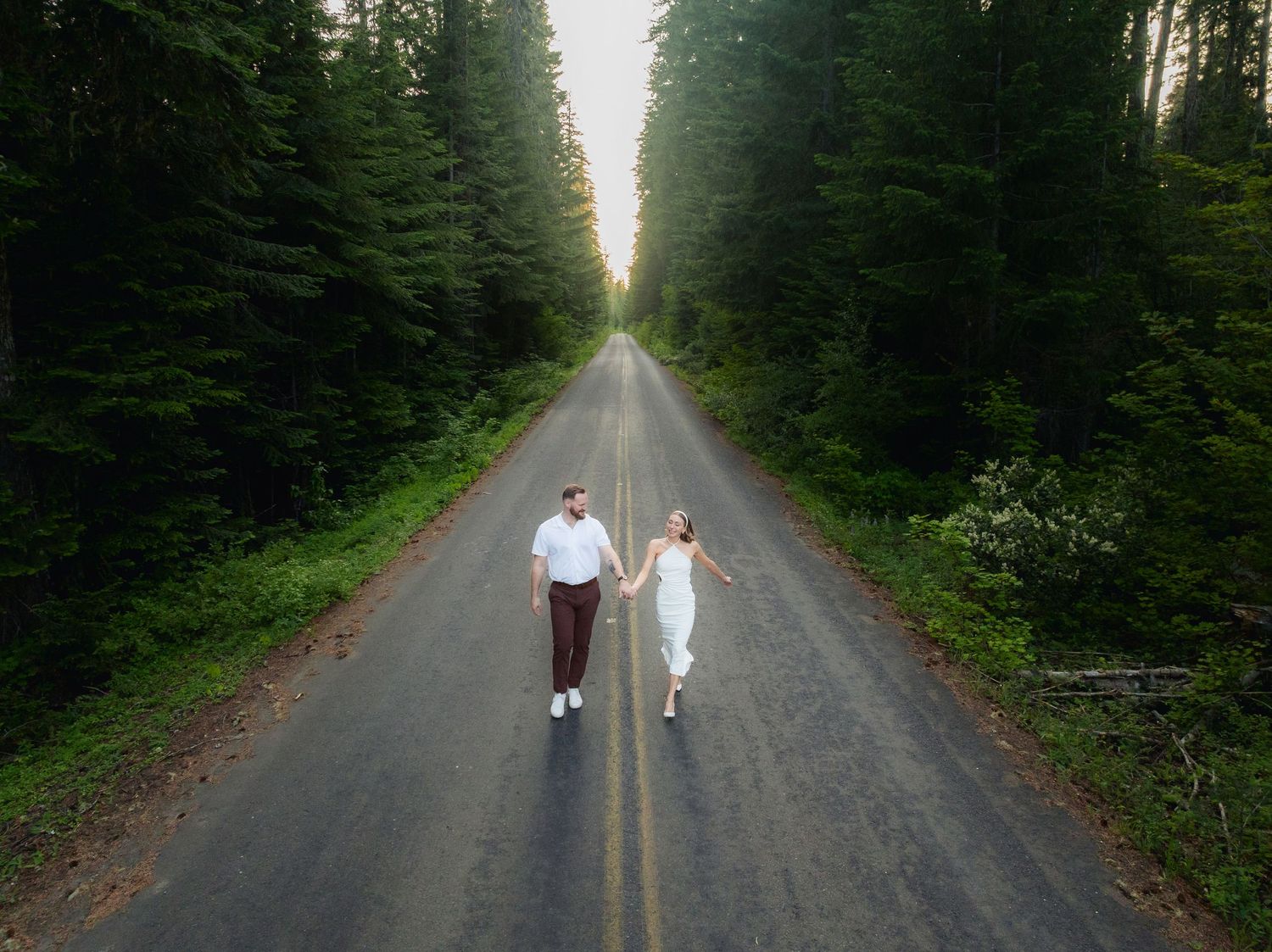 Couple holding hands walking on a tree-lined road in a lush green forest, aerial view of romantic outdoor photoshoot.
