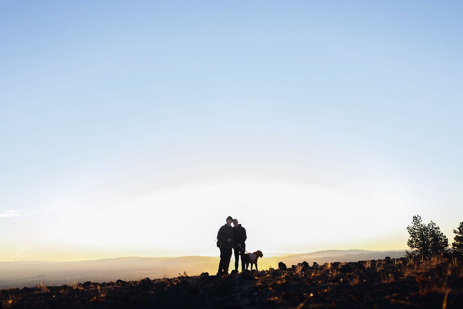 Silhouette of a couple embracing with their dog on a rocky hilltop at sunset, with a glowing horizon and distant mountains.