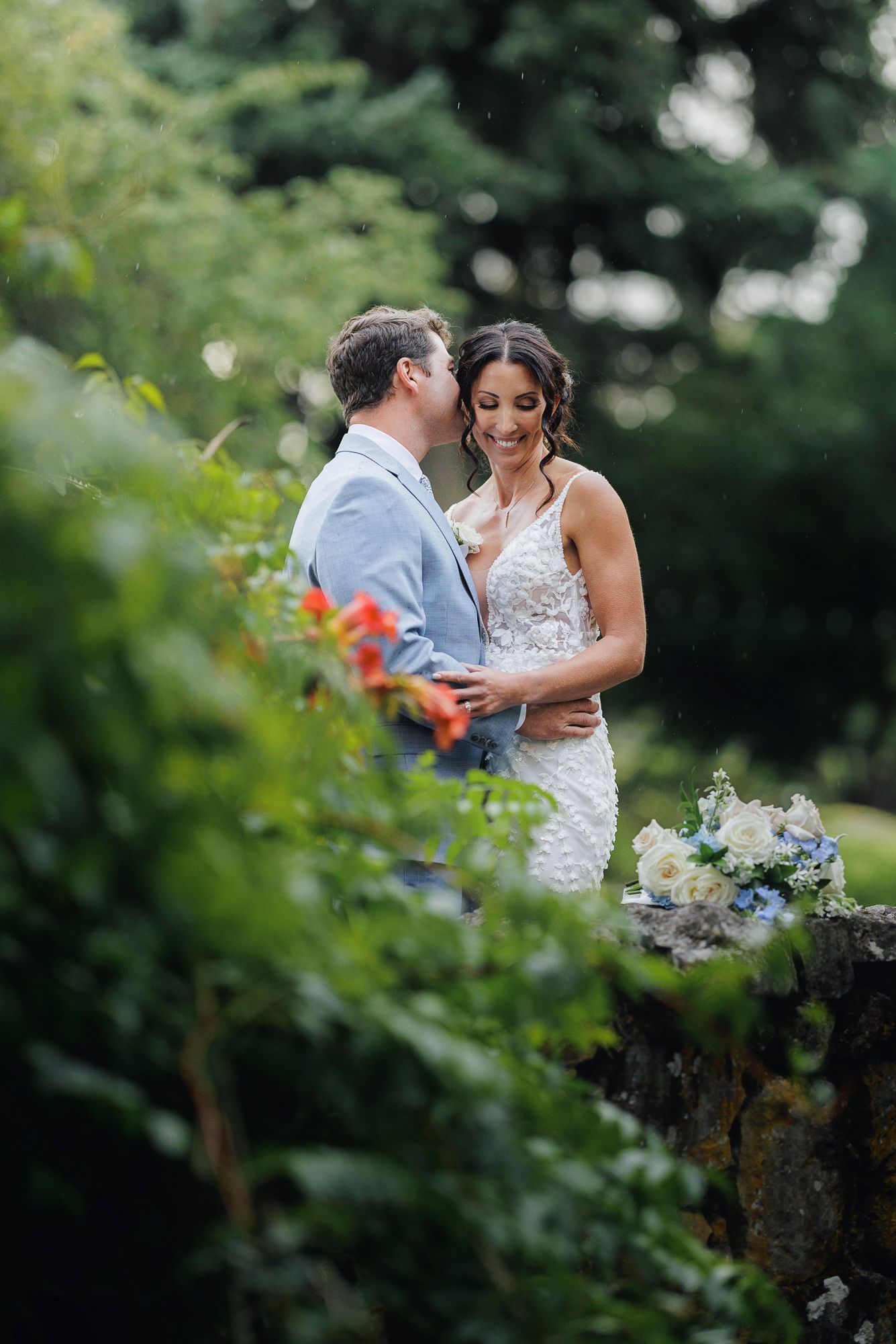 Romantic wedding couple embraces in a garden setting with soft natural lighting and blurred foliage.