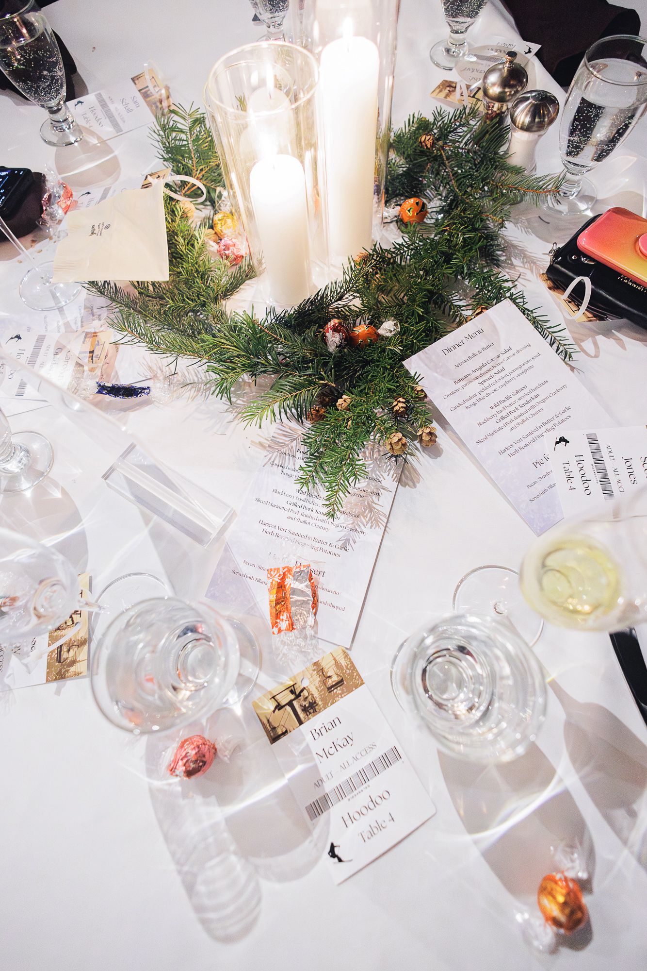 Wedding reception table settings with wine glasses, evergreen decor and menu cards on white tablecloths.