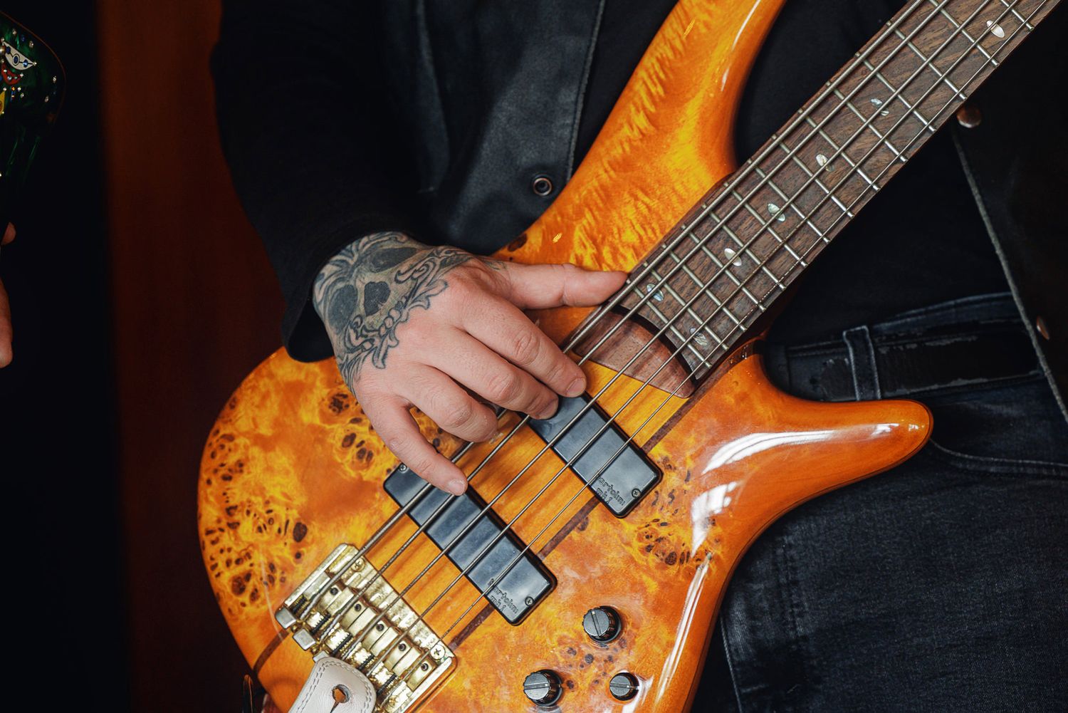 Tattooed hand playing an orange burst electric bass guitar, close-up shot of musician performing live.