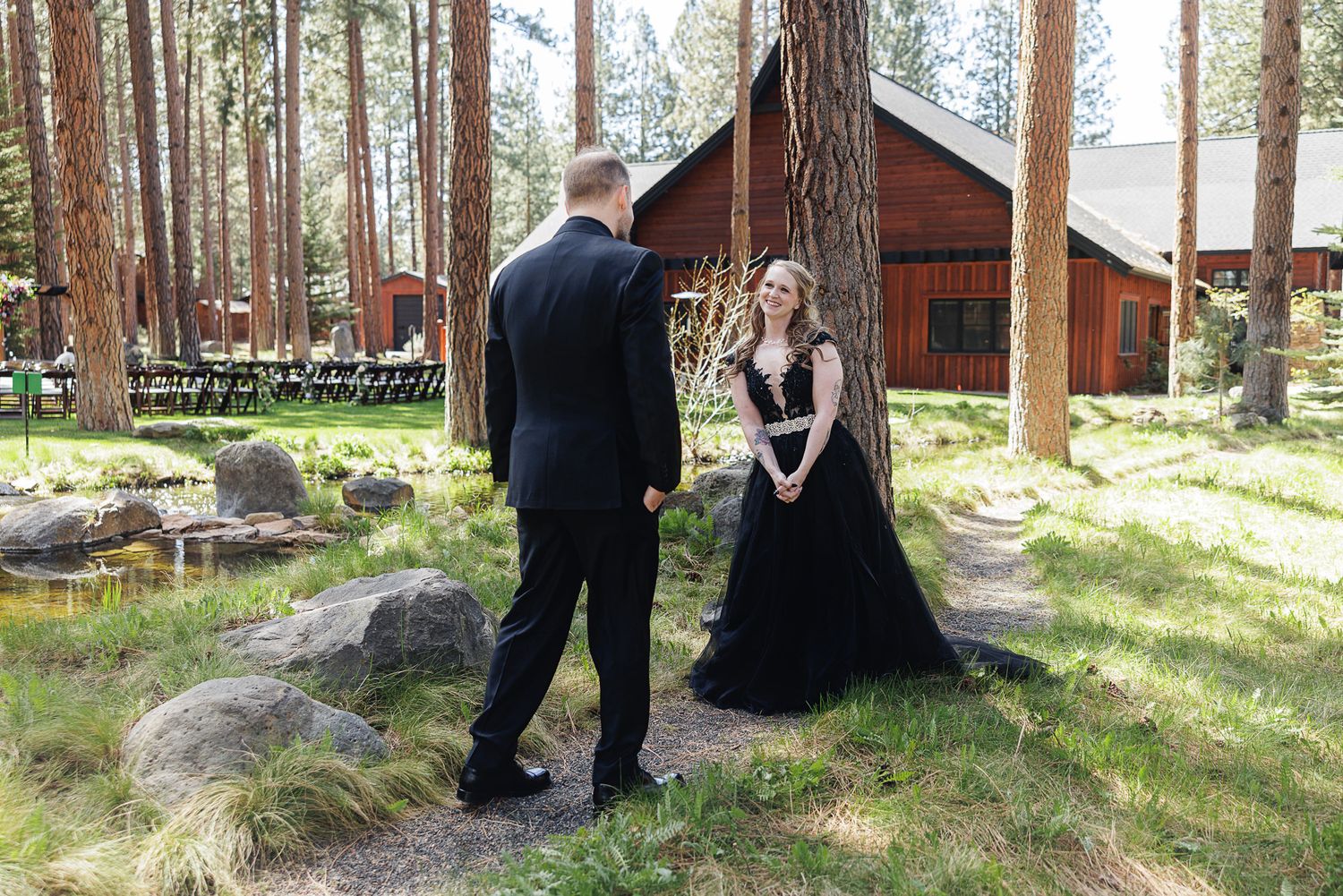 Couple shares first look moment in forest wedding venue, bride in black gown, groom in suit, rustic barn backdrop.