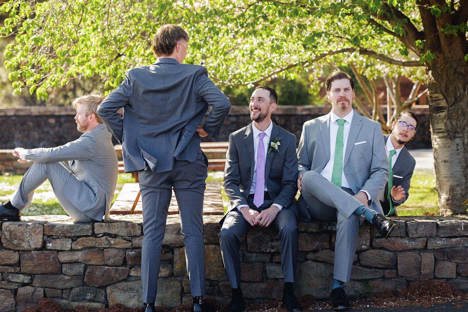 Groomsmen in gray suits with colorful ties pose playfully on a stone wall outdoors at a wedding celebration.