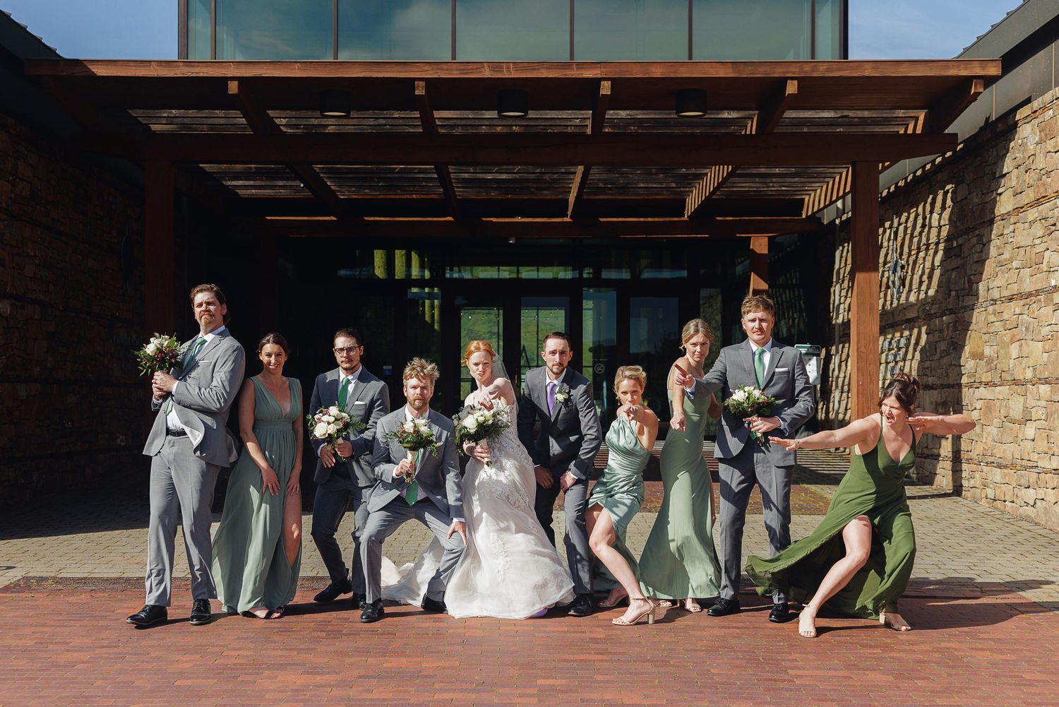 Wedding party in gray suits and sage green dresses posing joyfully outside a modern brick venue on a sunny day.