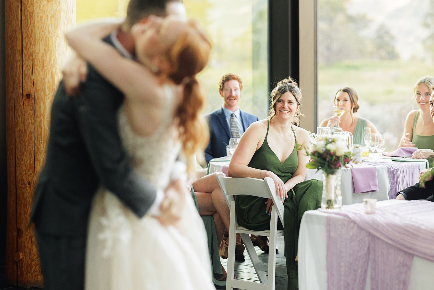 Bride and groom share first dance kiss while bridesmaid in green dress watches smiling at elegant wedding reception.