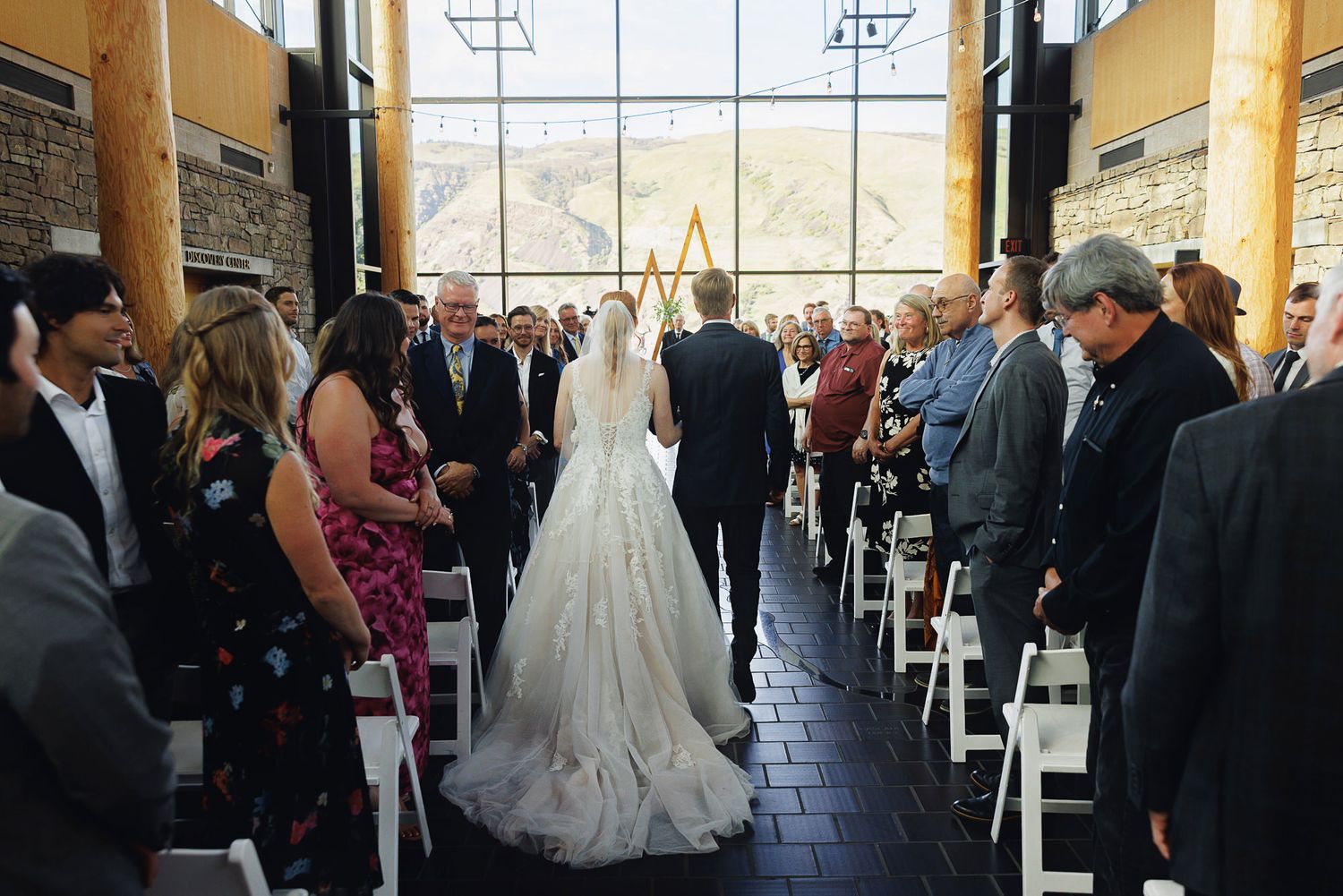 Bride walks down the aisle in a ballgown at an elegant mountain wedding venue with large windows and white chairs.