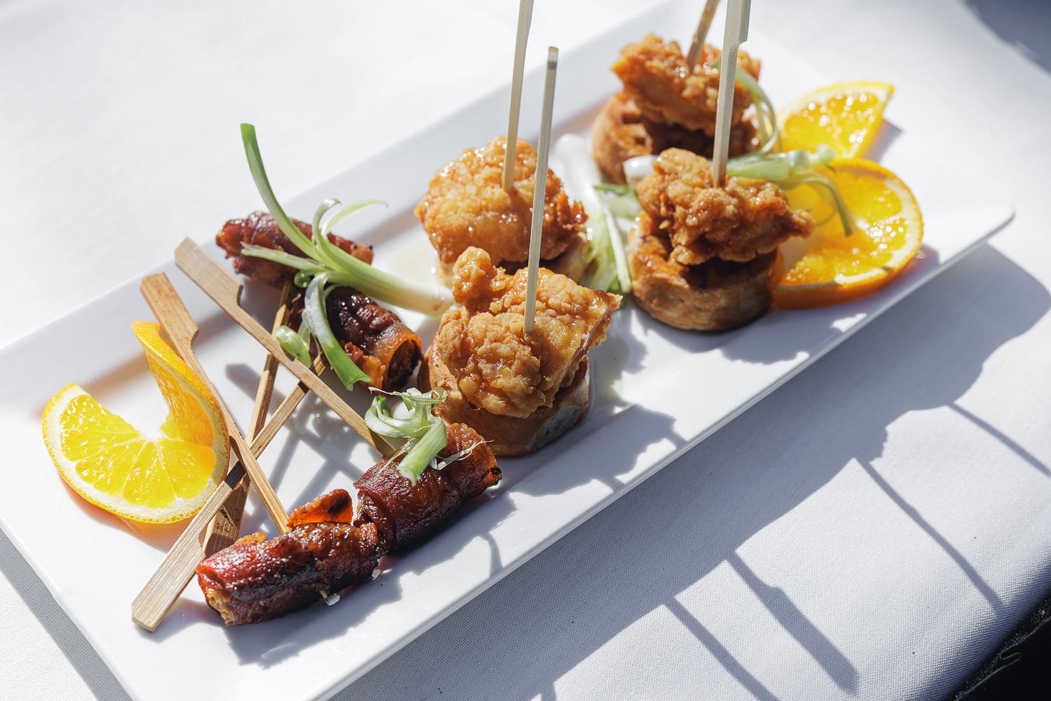 White plate with Asian appetizers: fried chicken bites, glazed skewers, green onions, and fresh orange slices.