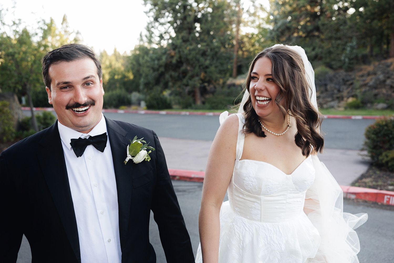 Happy bride and groom laughing together outdoors, wearing wedding attire including tuxedo and white bridal gown.